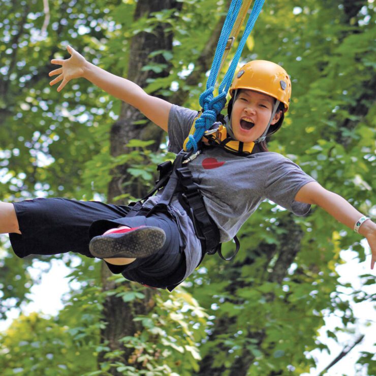 A woman ziplining amid trees wearing a yellow helmet and holding her arms out.