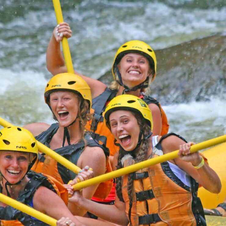 People in life vests and yellow helmets raising their paddles in a raft
